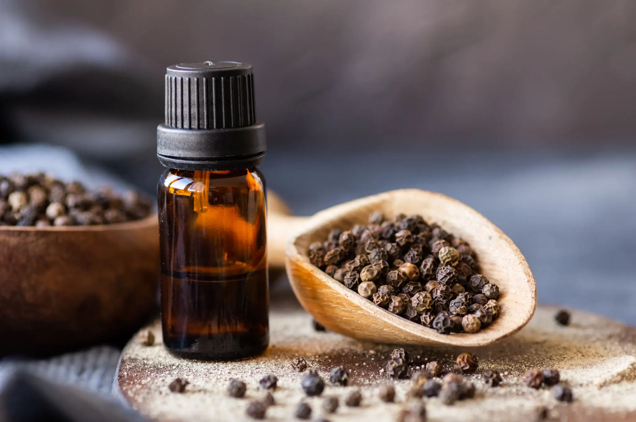 An amber bottle of black pepper essential oil beside a scoop of whole peppercorns
