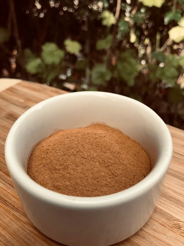 A white bowl of fine-ground Ceylon cinnamon on a bamboo board, with estate greenery behind.