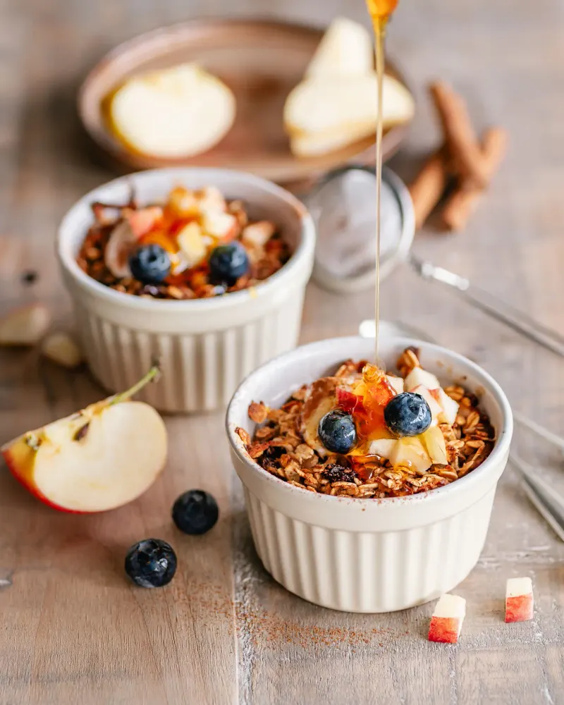 Two ramekins of baked oats with apple and blueberries, cinnamon quills alongside