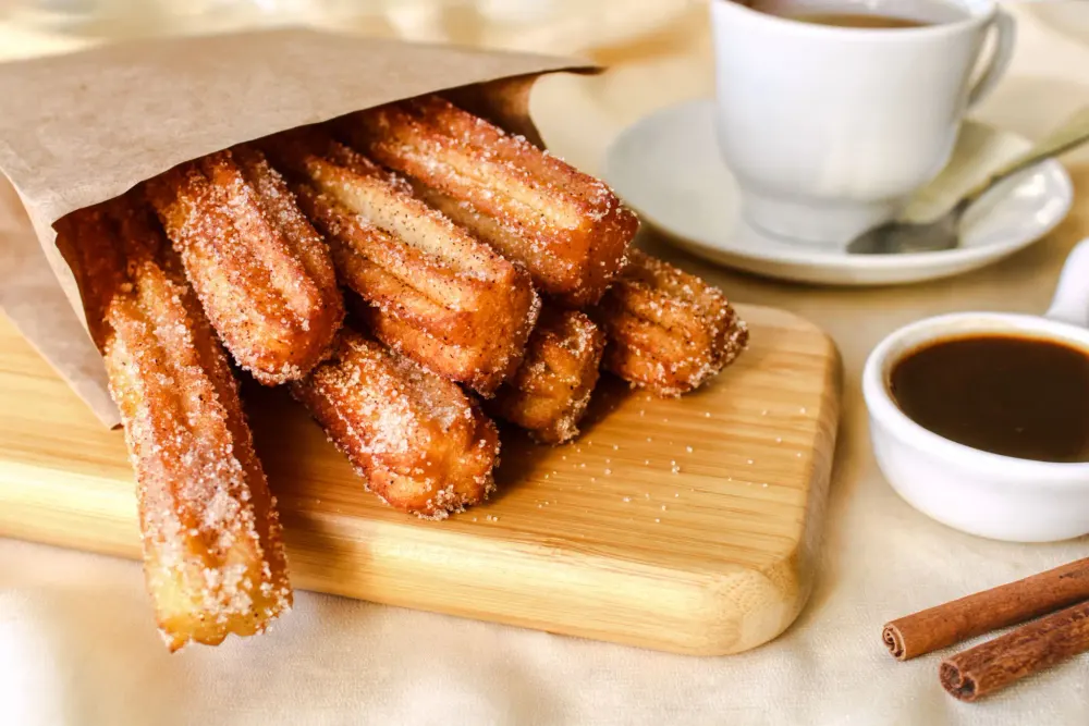 Churros in a paper cone on a board with a cup of coffee and a bowl of chocolate sauce