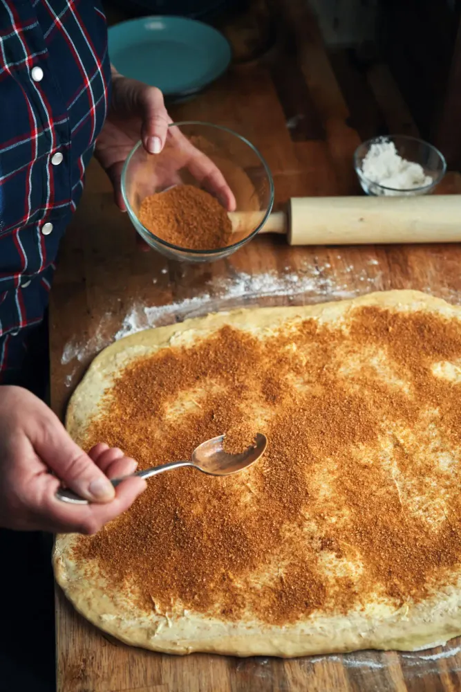 Cinnamon being sprinkled over rolled dough on a floured bench, a rolling pin alongside